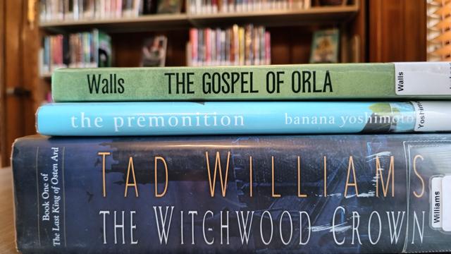 photo of three books in a stack, with library book shelves in the background. The Gospel of Orla by Eoghan Walls, the premonition by banana yoshimoto, and The Witchwood Crown by Tad Williams.