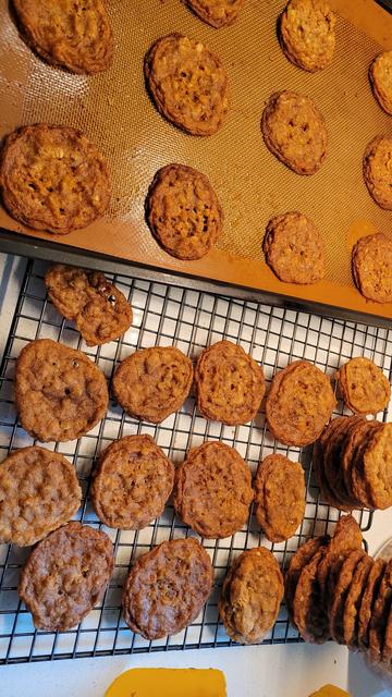 Photo of a pan of brown cookies with flakes of oat showing sitting on a baking rack that also holds stacks of the same cookies.
