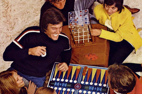 1971 A Gathering of Family or Friends Enjoying a Game Night

"This image shows a group of people engaged in what appears to be a board game.
The central focus is a wooden table with a game board that has several small pieces, suggesting it's a strategy game of some sort.
There are four individuals visible; two are seated at the table and two stand behind them.
All are smiling and seem to be enjoying themselves.
They are dressed in casual attire, and one person is holding a cup which might suggest they are drinking tea or another warm beverage.
The background shows a typical living room setting with a couch partially visible."