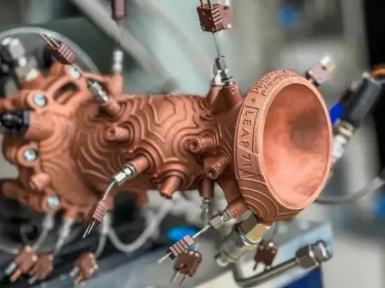 [A photo of a small rocket thruster, 3D-printed out of copper. Many sprockets and plugs are placed in an oddly haphazard manner, and the whole thing is ribbed and rippled in odd ways, concentric circles of ridges seeming to emanate from the sprocket attachment points. "LEAP 71" is engraved on the nozzle's rim).]