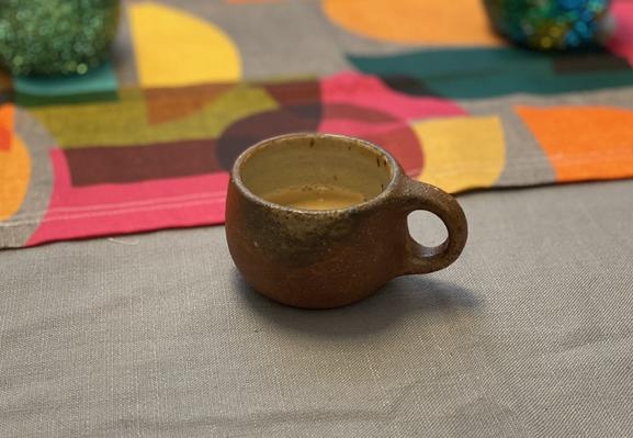 Repost of a mug shot from April, which features a tiny espresso cup in a rough reddish-brown glaze, on a table with a gray tablecloth and a colorful vintage-looking runner.  Don't let the inexplicable artistry of this shot discourage the more typical "my drink in my chaotic work space" photo that we all love to see!  mugs, mugs, mugs...