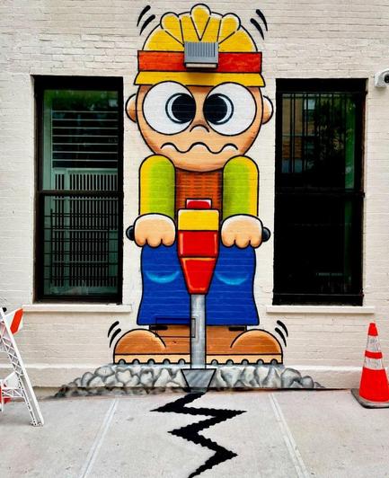 Streetartwall. A comic-style mural with an energetic construction worker was sprayed/painted on the outside wall of a building in New York. A construction worker with a yellow helmet and green/blue work clothes is now standing under a lamp on the wall, holding a jackhammer with both hands. His facial expression is very strained, but at least a (painted) crack can already be seen on the floor. The highlight of the small mural is the real wall light, which has been integrated into the painted helmet and is equipped with a motion sensor. So the little man lights up and works every time someone walks past