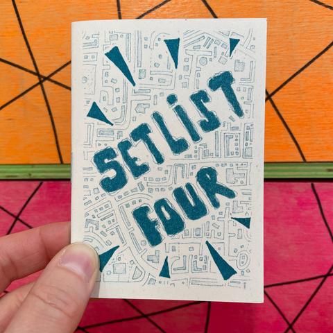It's another A7 zine held by a hand in front of a brightly-coloured painted wooden background (neon orange, green and pink with some black marker pen lines on it). This zine is Setrist Four, and the cover is just the title in hand-drawn text on a map-like background. The print is in teal ink.