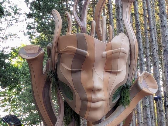 A large sculpture of a woman's face in mixed brown stripes blends into the trees.