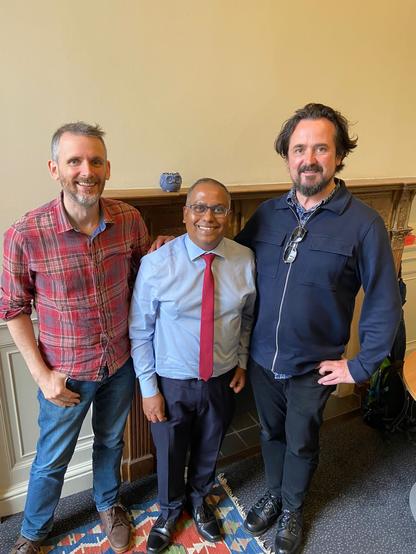 Colour image - three men standing in front of a wooden manetlpiece. All are smiling broadly. In the middle stands Ahmedul, the man of the moment, dressed in a pale blue shirt with red tie.