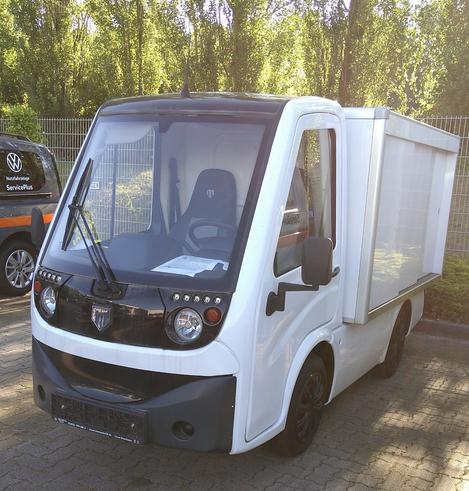 Ein kleiner Elektrotransporter mit sehr kompaktem Führerhaus und dahinter eine rechteckige Box mit Schiebetüren. Wie ein viel zu heiß gewaschener und geschrumpfter Transporter. Die Farbe ist weiß, Stoßfänger, Spiegel, Türgriffe und ein paar Anbauteile schwarz als Kontrastfarbe.