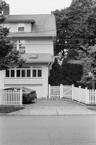 A Honda EV sits in a driveway behind a white picket fence with a three story home behind it.