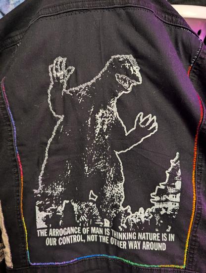 Photo of a black patch with a white line image against black. It shows Godzilla about to stomp a building with text: "The arrogance of man is thinking nature is in our control, not the other way around.

The patch is sewn to the jacket in a square of rainbow colors. Godzilla's outline is half embroidered with silver grey thread.

Rawr.