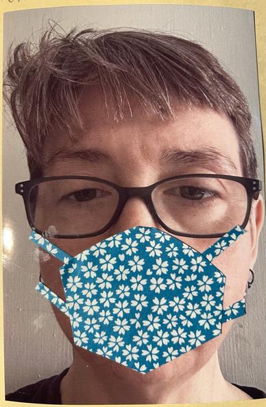 Collage in my sketchbook. Photo of a white Roman with short brown hair and rectangular glasses with dark frames. Over her face is a mask like we’ve all been wearing the last few years made of blue origami paper with white flowers.