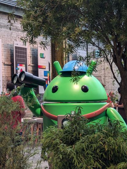Inside the venue at I/O Connect Berlin: A statue of a green Android with a blue hat, holds a pair of binoculars while standing behind some trees.