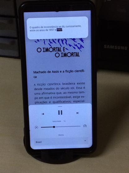 Smartphone, rodando o app BibliON (da Biblioteca Digital Gratuita do Governo de SP). O e-book é "O Imortal e O Imortal", de Machado de Assis. A tela mostra o modo de Texto para Fala, no fundo, a página do livro, no topo, a palavra que está sendo falada no momento, no rodapé os controles do reprodutor de áudio, com opção de retrocesso, pausa, avanço e controle de velocidade.