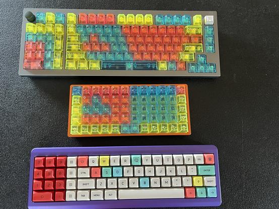 Three colorful mechanical keyboards on a dark surface. The keyboards feature vibrant, multi-colored keycaps in red, yellow, blue, teal, and purple.