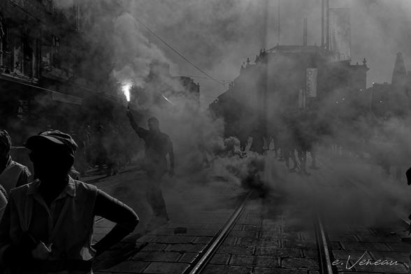 In an urban setting, during a popular protest against government policy, a man holds a smoke bomb like a torch in the middle of a crowd whose silhouettes can be seen in an atmosphere saturated with tear gas.