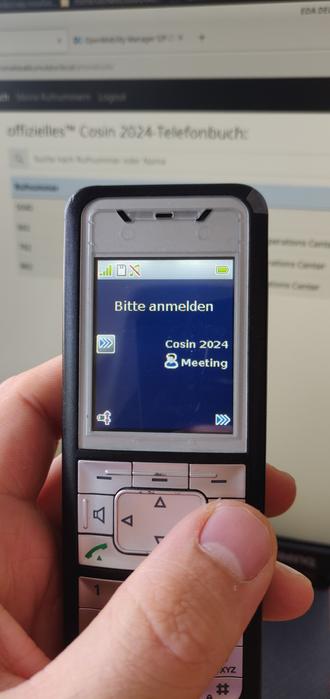 Ein Mitel DECT-Telefon, was Cosin 2024 anzeigt. Im Hintergrund steht ein Monitor mit dem Telefonbuch