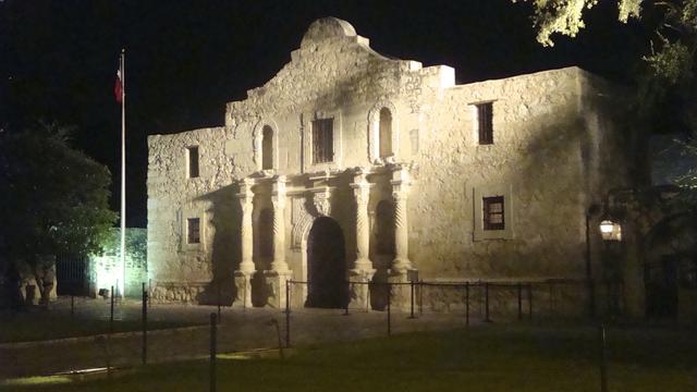 Historisches Steingebäude (The Alamo), nachts beleuchtet, mit einem Fahnenmast und Bäumen in der Nähe.
