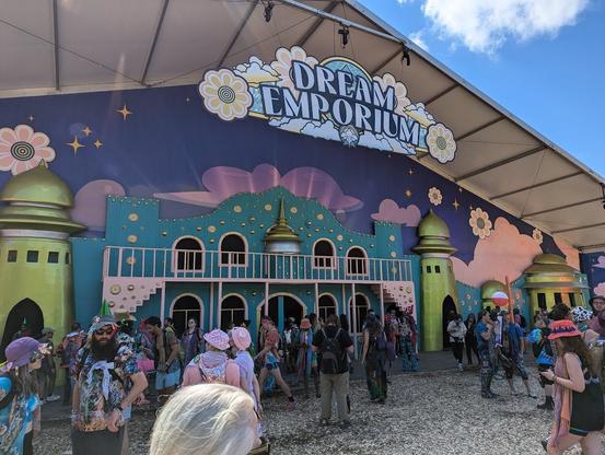 A brightly colored facide of a building with golden spire type towers, bright clouds, golden shields and many windows.  In front of the building are multiple festival-goers.
