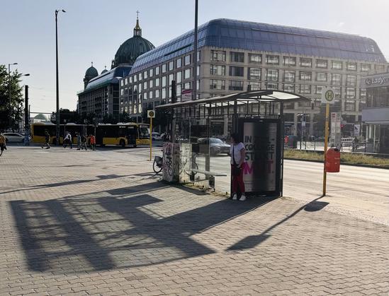 Urban street scene with a bus stop, pedestrians, and yellow buses. A large building with a dome is in the background In the forground a person in the shadow at the back of a Bus stopp.