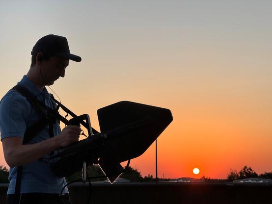 Wolkenloser Abendhimmel mit untergehender Sonne und Abendrot. Im Vordergrund ist der Schattenriss einer Person zu erkennen, die in beiden Händen Messgeräte hält.