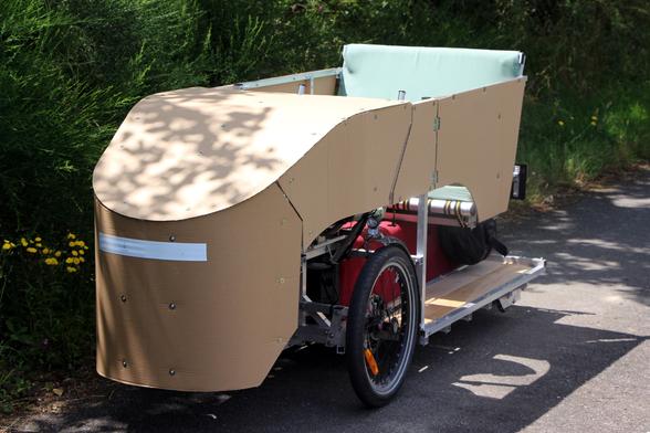 Photo of the same vehicle, but in real world this time. It is empty and stationed on the side of a small countryside road. On this version, the enclosure is prototyped out of cardboard.