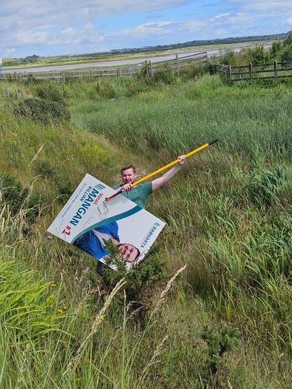 A photo of Killian holding a pole with a hook in one of his local election poster retrieved from a grassy ditch.