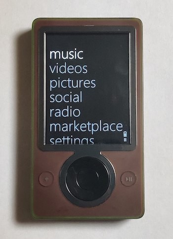 A brown Zune (brown casing, black features) that's on, with its menu open. Categories from top to bottom in the list are music, videos, pictures, social, radio, marketplace, settings, and a partially full battery indicator at the bottom.