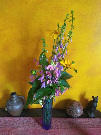 Orange false sunflowers, purple creeping bellflower, pink wild sweet pea in a blue glass bud vase sitting on a brown table with red runner cloth in front of an orange color washed wall. The vase is flanked by a brown pottery jug, a brown ceramic ball and a dark brown Bast statuette.