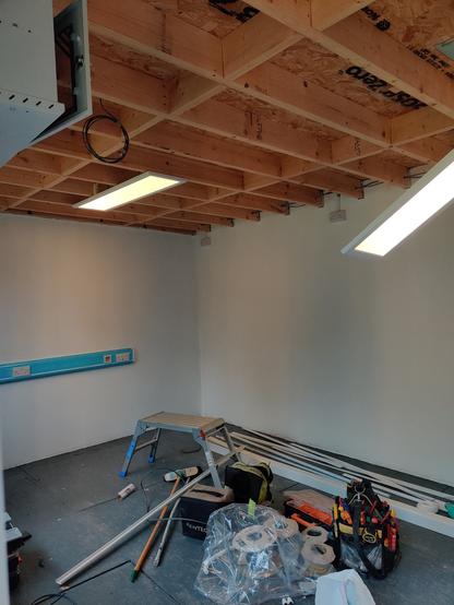 inside of garden office with messy electricians tools and supplies over the floor. on the ceiling 2 panel lights are glowing. one fixed to the ceiling, the other still suspended by metal rope fixings.