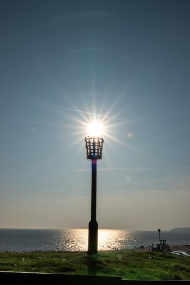 An empty fire beacon basket on a tall pole has the sun shining just above/behind it. The beacon sits on a grassy area with the wide sea behind.