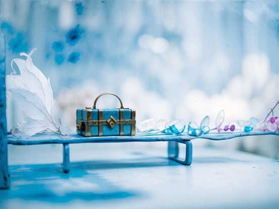 Fairy-like setting in blue hues, of a miniature bench made of found material, holding a small blue chest with gold accents, white butterfly wings, and translucent beads arranged delicately, against a softly blurred background.