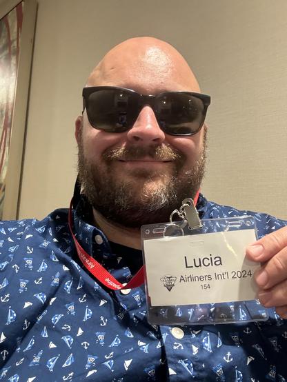 Bald guy with a beard wearing large sunglasses and a fun shirt shows off a convention badge which belongs to Lucia which will be used to crash a party he wasn’t invited to.