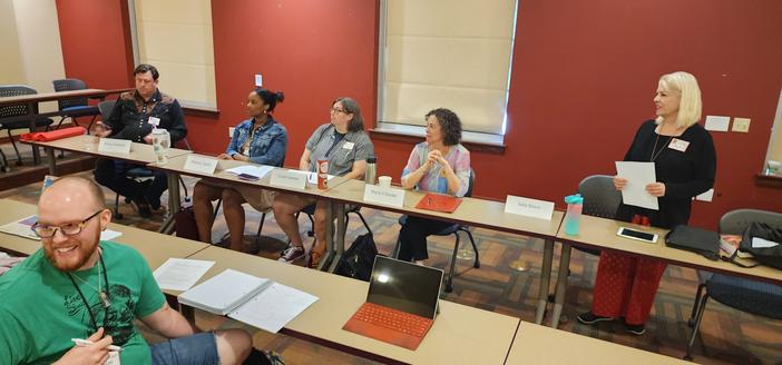 Five person panel sitting at a long table, looking at each other and discussing. Person in front row turned toward the camera, smiling.