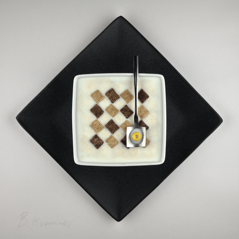 Seen from above, in a square image,on a white background sits a black plate with the points towards the edges. On that sits a smaller white bowl, aligned to the frame. The bowl is filled with milk, and has a checker board pattern of Chex cereal pieces. The rectangular pieces are rotated to match the plate, and alternate between light and dark colors. In top of the cereal site a spoon, with all the rounded edges squared off. In the spoon is a small pool of milk, and a single yellow round Fruit Loop.