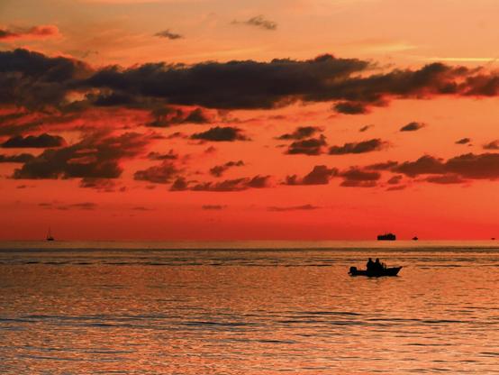 A beautiful sunset over the ocean with silhouettes of boats and scattered clouds in the sky.