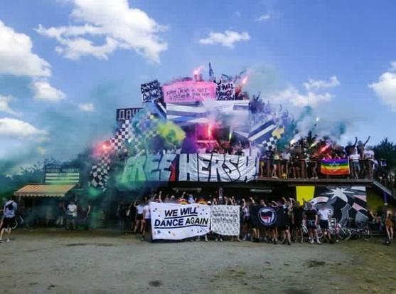 Group of people gathered outside a building holding banners and signs with colorful smoke and bright lights.