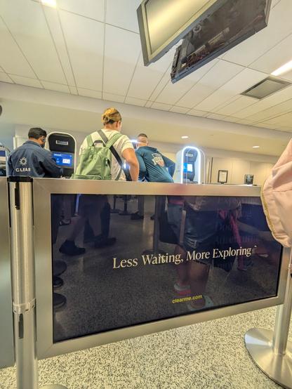 Travelers queue up to clear (tm) booths at a US airport to get through the security line faster. A banner advertisement on the divider between the Clear and regular customers reads "Less waiting. More exploring."