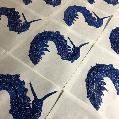 A collection of prints on 8” x 8” square of white washi paper with a blue S-shaped velvet worm, resembling a many-legged caterpillar with two antennas and a pattern of spots. Along its length there’s a central darker blue stripe. There look to be a grid of 12 prints.