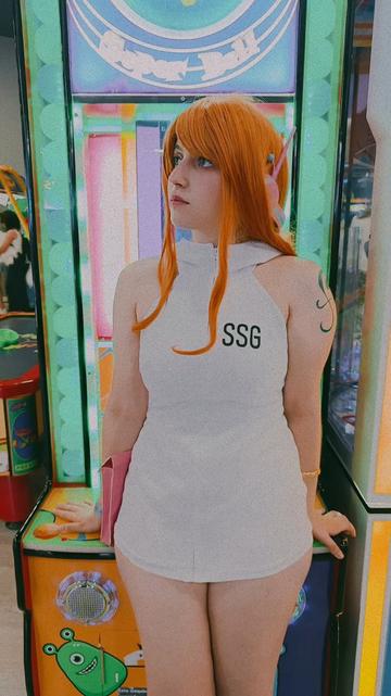 Aparezco con el cosplay de Nami apoyada en una máquina de juegos con luces verdes.