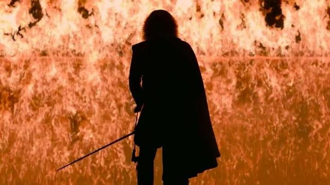 A man standing in profile against a huge wall of flame. He's wearing a cape and has a sword