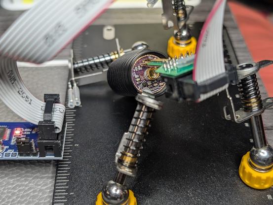 Picture of a flashlight head being held in place by clips on an omnifixo "third hand" device. Another clip on the device is holding a probe which is pressing pogo pins onto flashing pads inside the flashlight head.

A USBASP device is visible on the left. A ribbon cable connects the USBASP device to the probe.