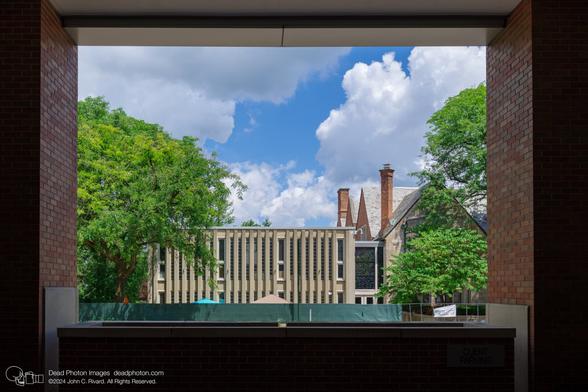 Brick columns and a limestone planter frame a building with vertical concrete columns and glass-block. It is connected by a stain-glassed bridge to a gothic limestone and brick building with peaked slate roofs and chimneys. Above are clouds and a blue sky.