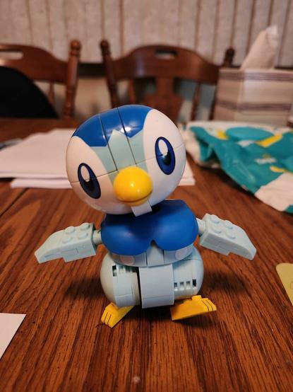 Photo. Piplup, a small penguin-like Pokémon, is recreated in not-Legos and stands on a cluttered wooden tabletop. The figure is posed to appear as if waddling toward the camera.