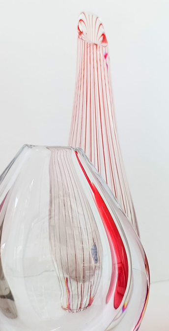 eine kleine dicke und eine hohe schmale Glasvase. Die hohe Vase steht hinter der dicken und hat feine senkrechte rote Streifen. Ihr unteres Ende wird durch das Glas der davonstehlenden Vase verzerrt