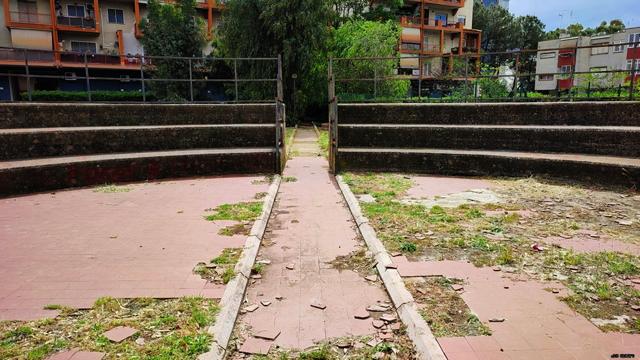 Farbfoto eines Amphitheaters in einer Wohnanlage in Rom Süd, das vermutlich für soziale Veranstaltungen der Bewohner und Anwohner vorgesehen war und seit Jahen ohne Nutzung vor sich hin verfällt. Im Vordergrund ein Teil des gefliesten Bodenrunds mit Gang in der Mitte, dahinter links und rechts eine Tribüne, im Hintergrund Wohnblocks im Stil des Brutalismus.