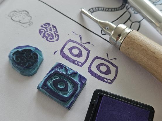 Foto eines Skizzenbuchs mit einigen Stempelabdrücken darin. Die zugehörigen Stempel, ein violettes Stempelkissen sowie Schneidewerkzeuge liegen daneben auf dem Buch.