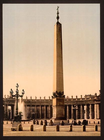 The image is a vintage photograph capturing the grandeur of St. Peter's Square in Rome, Italy, likely taken during the late 19th or early 20th century. The square features an impressive obelisk as its centerpiece, which stands tall on a stone pedestal. Flanking the obelisk are two large fountains with statues and other architectural details that contribute to the overall elegance of the scene. The photograph offers a view of the square from one end to another, showcasing the layout and design elements of this significant landmark.