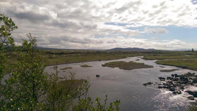 A river flowing through a grassy landscape with some rocks, surrounded by green vegetation and under a cloudy sky. Distant mountains are visible on the horizon.