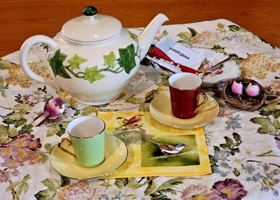 A tea tableau on a dark yellow, green, mauve, and cream tablecloth, with an ivy-patterned teapot & little magenta mushroom birds surrounding demitasse cups & saucers.