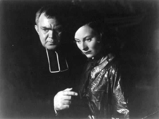The image is a vintage black and white photograph featuring two individuals. On the left side, there's an older man dressed in what appears to be clerical attire, including a collar that suggests he might be a priest or religious figure. His facial expression is serious, and his body language seems introspective or contemplative.
On the right side of the image is a woman who seems to be in her mid-adulthood. She has short hair and is wearing what looks like a dark top with some light embellishments on it. Her expression is one of concern, and she is looking intently at the man, perhaps as if they are engaged in an important conversation or he's sharing something significant with her.
The background is dark and out of focus, which puts more emphasis on their expressions and interaction. The image has a vintage feel to it, possibly indicating it was taken several decades ago, and the quality suggests it could be from a film still or an older photograph meant for archival purposes. There are no visible texts in the image to provide additional context about the individuals or the situation depicted.