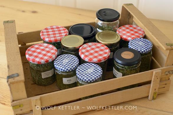 The photograph show jars with tops of different colours and patterns standing in a wooden crate on the wooden surface of a rustic table. The jars have white labels attached to their sides identifying the contents. They are filled with dried herbs.