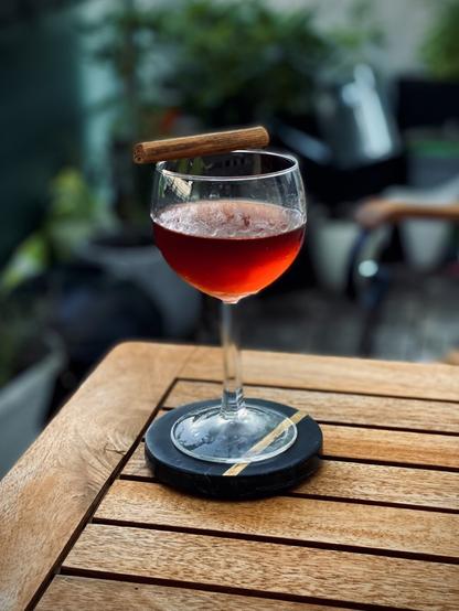 ruby red cocktail in a large round coupe glass with cinnamon stick garnish on a wooden table with plants in the background